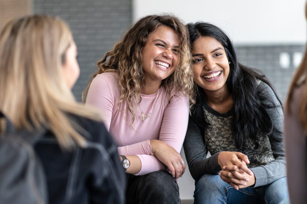Two female young adults sit closely together, with their heads resting against each others, during a group therapy session. One is of African decent and the other is of Indian decent. Bother are dressed casually and are sitting among their multi-ethnic peers with smiles on their faces.